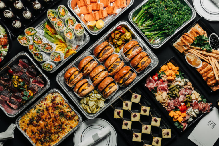 Overhead view of catering trays with sushi, sandwiches, and skewers for a rotating office menu